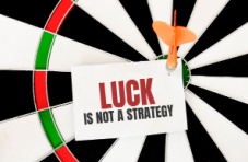An image of a dartboard with a dart pinning down a posted note reading luck is not a strategy on a white background 