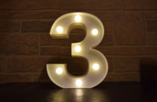An illuminated number three with light bulbs on a table against a dark brick wall