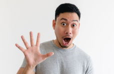 A photo of a man with a surpised look on his face making the number five with one hand on a white background