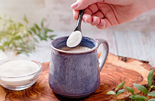 A photo of a woman’s hand adding a teaspoon of collagen to a mug of coffee 