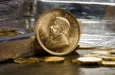 A photograph of a South African Krugerrand gold coin standing upright among scattered gold coins on a metallic background