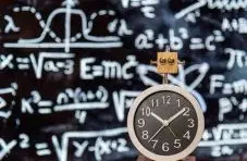A photograph of a clock with a tiny wooden robot on top against a dark chalkboard with math formulas background