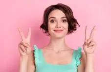 A close-up photo of a smiling short-haired woman in mint top making double peace signs on pink