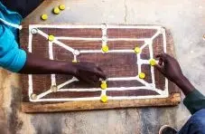 A photograph of two people playing Morabaraba using bottle caps on a homemade wooden board on a concrete background