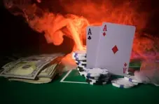 An image of two aces, casino chips & dollar bills on a green felt table with smoke, flames in the background