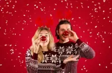 A photo of two young people wearing faux reindeer antlers and holding Xmas baubles to their nose