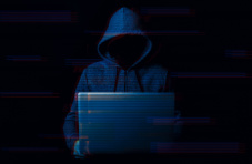A dark image of a hooded person sitting at a laptop