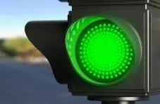 A close-up photograph of a glowing green traffic light against a blurred street and blue sky background