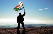 A photograph of a person's silhouette waving a South African flag on a hilltop with mountains and blue sky background