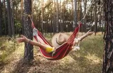 A photo of a person relaxing in a colourful hammock tied between trees in a sunlit forest, with arms stretched out in a carefree