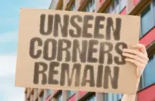 An illustration of hands holding a cardboard sign reading “Unseen Corners Remain” in front of an urban building background