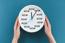 Female hands holding a white clock with the word 'NOW' in place of each hour, against a blue background