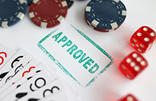 An image of a green ‘approved’ stamp with playing cards, casino chips and a pair of red dice on a white surface