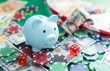 A photograph of a blue piggy bank surrounded by casino chips, dice and dollar bills on a green background