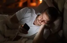 An adult man lying in bed at night, illuminated by the glow of his smartphone while looking at the screen in a dimly lit bedroom