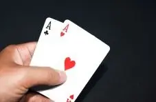 A photograph of a hand holding two ace playing cards showing clubs and hearts on a dark background