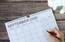 A photo of a spiral-bound September 2026 calendar with a hand holding a pen on a rustic wood table