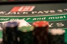 A close-up photograph of a blackjack table felt showing the soft 17 rule text with blurred chips and cards on a green background