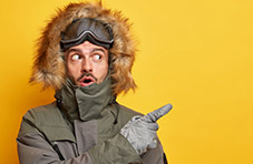 A photo of man dressed for winter weather with a surprised expression, pointing over his left shoulder on an orange background
