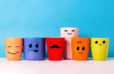 A photo of 6 different coloured and sized coffee mugs with funny faces drawn in black marker