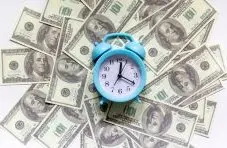 A photograph of a blue alarm clock placed on a spread of US hundred dollar bills on a white background