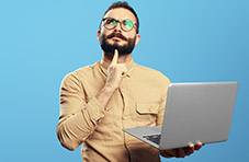 A photo of a bearded man deep in thought holding an open laptop in one hand on a blue background
