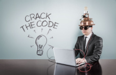 A man at a table in front of a laptop with a vintage gadget on his head with ‘crack the code’ text on a light background 