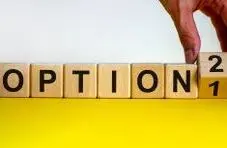 A photograph of a hand flipping a wooden cube from "Option 1" to "Option 2" on a yellow and white background