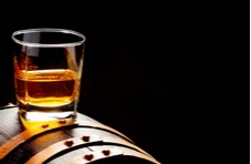 A image of a glass tumbler half filled with whiskey, balancing on the side of a whisky cask on a dark background