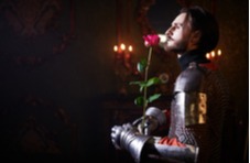 A photographic image of a handsome knight in armour holding a long stemmed red rose on a black candle-lit backdrop