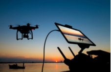 A silhouette of a military drone at sunset operated by a man’s hands via a tablet device