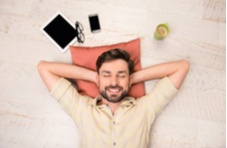 A smiling man sleeping on his back on a wooden floor with a notebook, mobile phone, mug of coffee and spectacles 